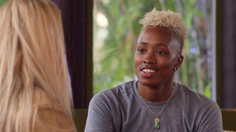 Mal Wright with short blonde hair in a conversation, wearing a gray top and a necklace, indoors