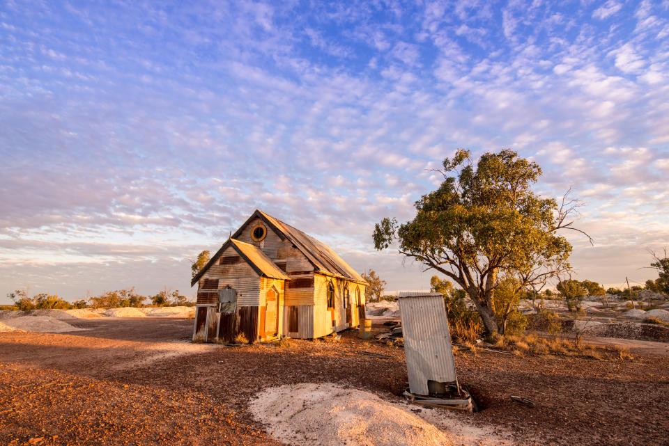 Rusty old corrugated iron church below mackerel sky of glowing cirrocumulus clouds at dusk. The church, once part of a film set, sits deserted on the dry opal fields at Lightning Ridge, a small town in northern New South Wales, Australia. A small corrugated metal dunny (toilet) stands at right.