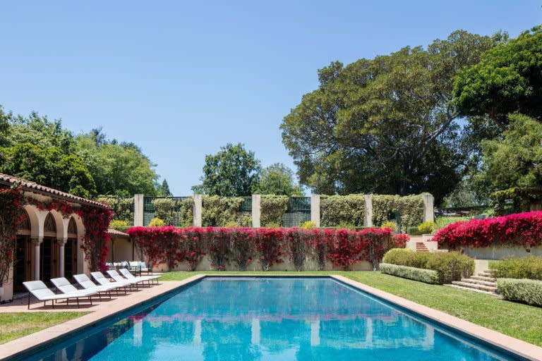 This shows Owlwood Estate's outdoor pool. It's a sunny day. There are sunbeds lined up on the left side of the pool, and pink flowers line the exterior of the pool. Source: Compass