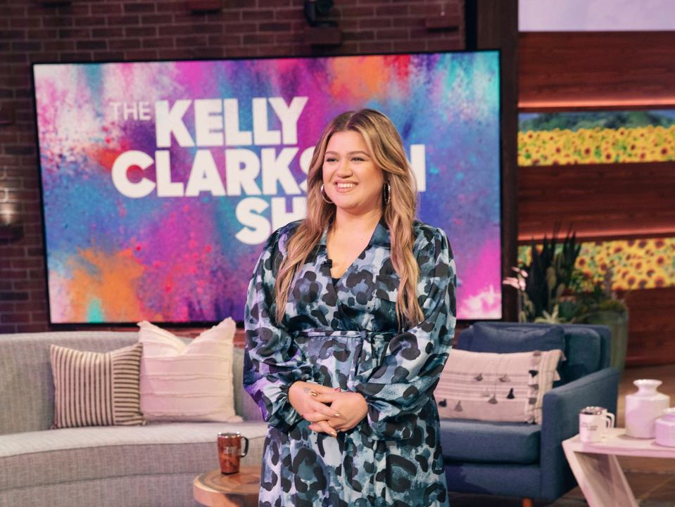 Kelly smiling in a blue dress in front of a Kelly Clarkson backdrop and set.