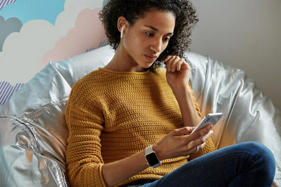 A young woman sitting down wearing AirPods and looking at an iPhone.