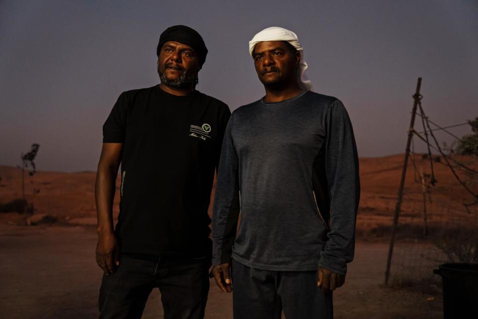 Two men in dark clothing, one wearing a white headdress, stand in a desert setting