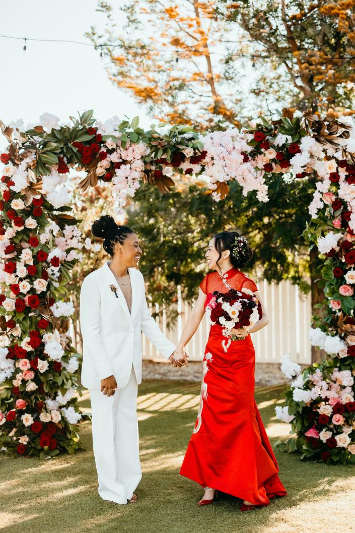 Two brides hold hands and smile at each other in front of a floral arch.