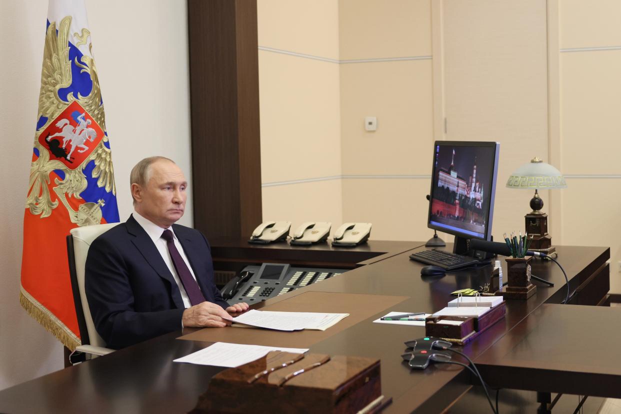 Vladimir Putin sits at a large desk with many phones and a flat screen.