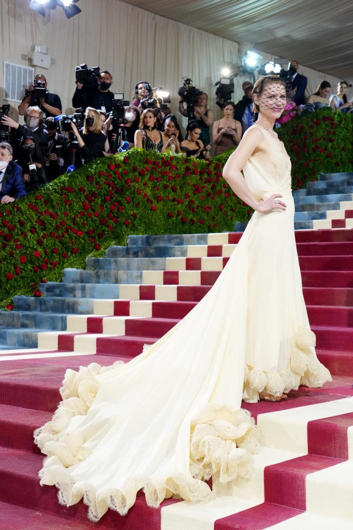 Claire on the red carpet dress in an off-white yellow-ivory dress with a long dress flowing down the steps. The top has a lace bodice and thin lace straps and the solid fabric on the skirt ends with a train with yellow ruffles. She's wearing a black birdcage headpiece over her face.