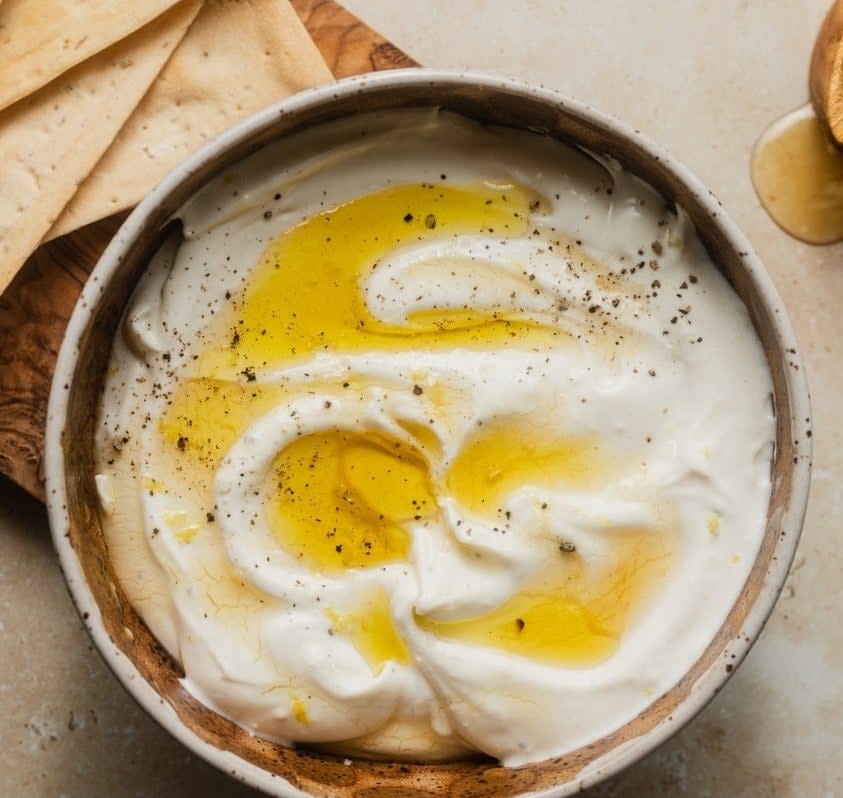 A bowl of creamy dip topped with a drizzle of olive oil and sprinkled with black pepper, accompanied by crackers on the side