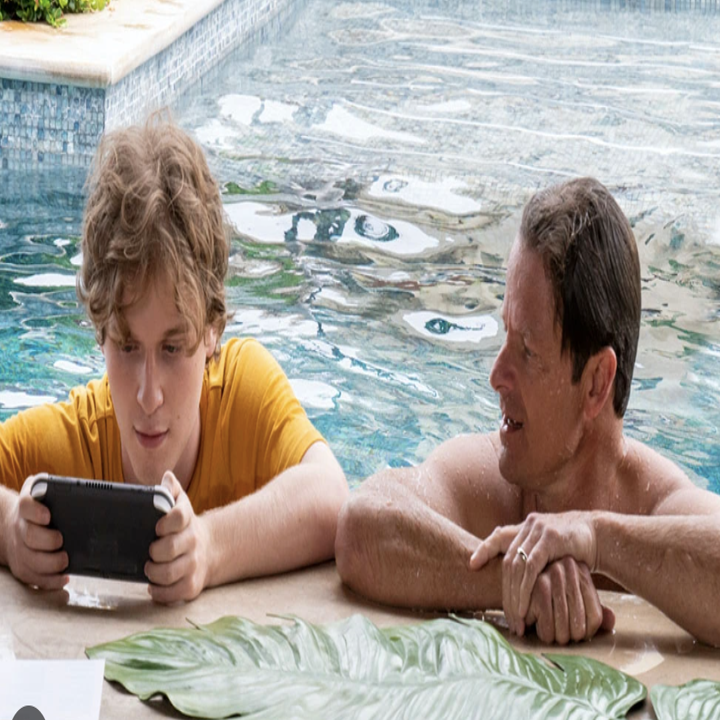 A kid plays video games in the pool with his father looking at him
