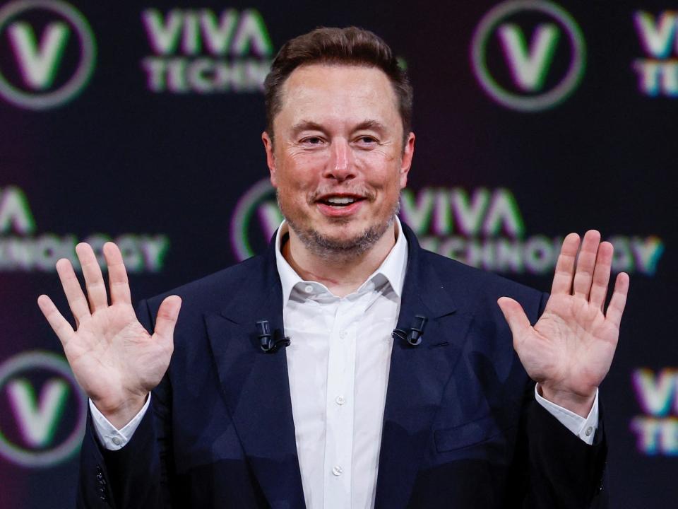 elon musk holds his hands up palms out smiling wearing a suit in front of a wall with the logo viva technology