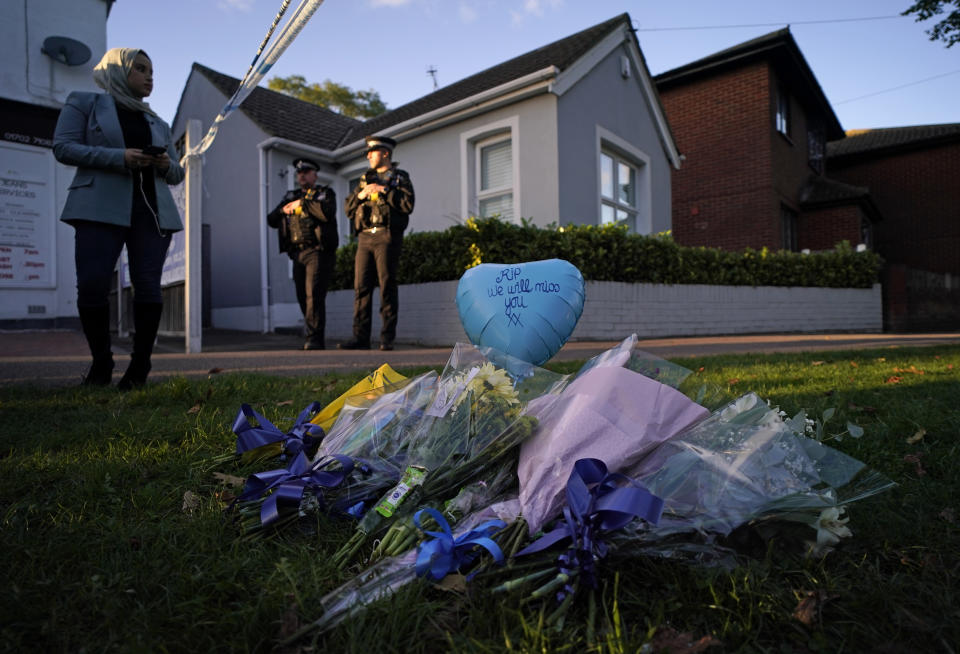 Floral tributes can be seen as police officers block one of the roads leading to the Belfairs Methodist Church in Eastwood Road North, where British Conservative lawmaker David Amess has died after being stabbed at a constituency surgery, in Leigh-on-Sea, Essex, England, Friday, Oct. 15, 2021. Police gave no immediate details on the motive for the killing of 69-year-old Conservative lawmaker Amess and did not identify the suspect, who was being held on suspicion of murder. (AP Photo/Alberto Pezzali)