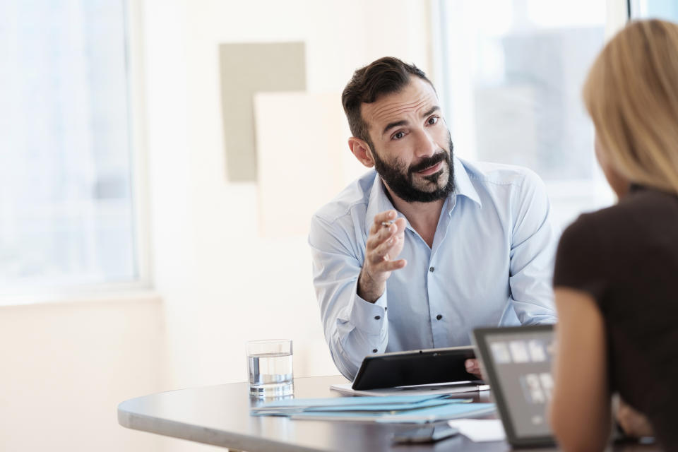 A man in a business meeting gestures while speaking to a woman, who is seen from behind, in a bright office setting