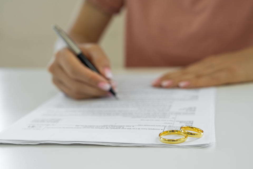 A person signs a document on a table with two gold rings placed nearby