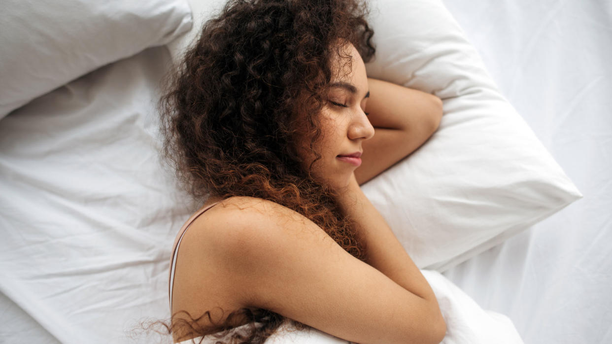  Woman with black curly hair sleeps on a white bed. 