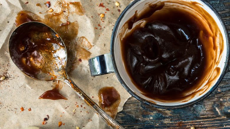 Cup of barbecue sauce next to metal spoon and chili flakes on parchment