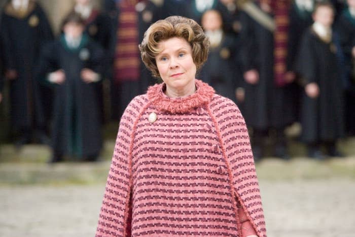 Imelda Staunton as Dolores Umbridge in a formal setting, wearing a patterned shawl. Background has a group of students in black uniforms and striped scarves