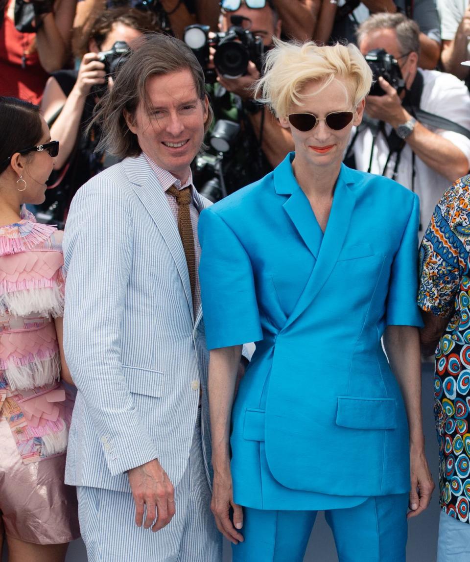 wes anderson, wearing a light blue suit, smiles next to tilda swinton, who wears a blue pantsuit and sunglasses