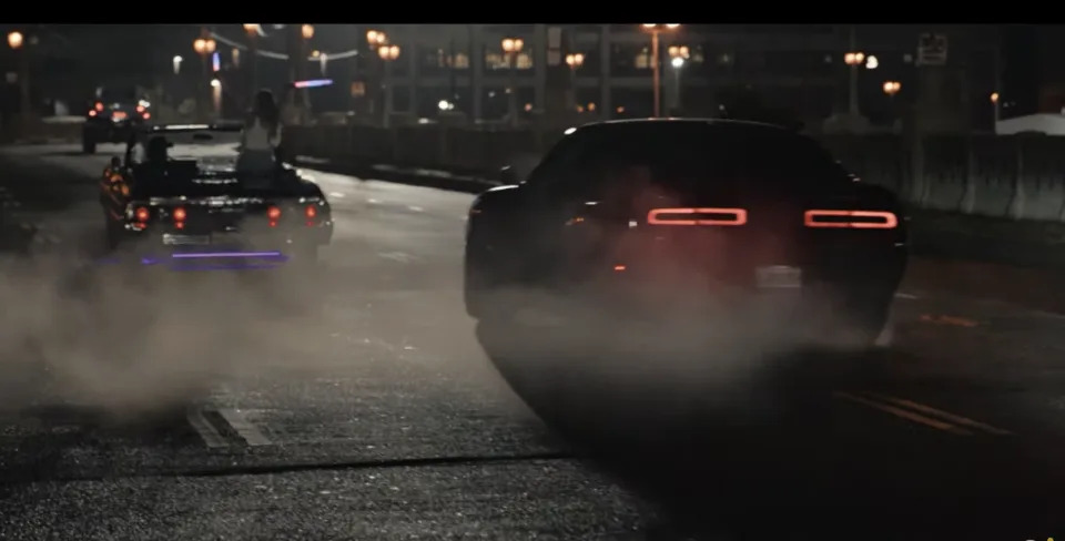 Two cars, one vintage with passengers and one modern, seen drifting on a city street at night amidst smoke clouds