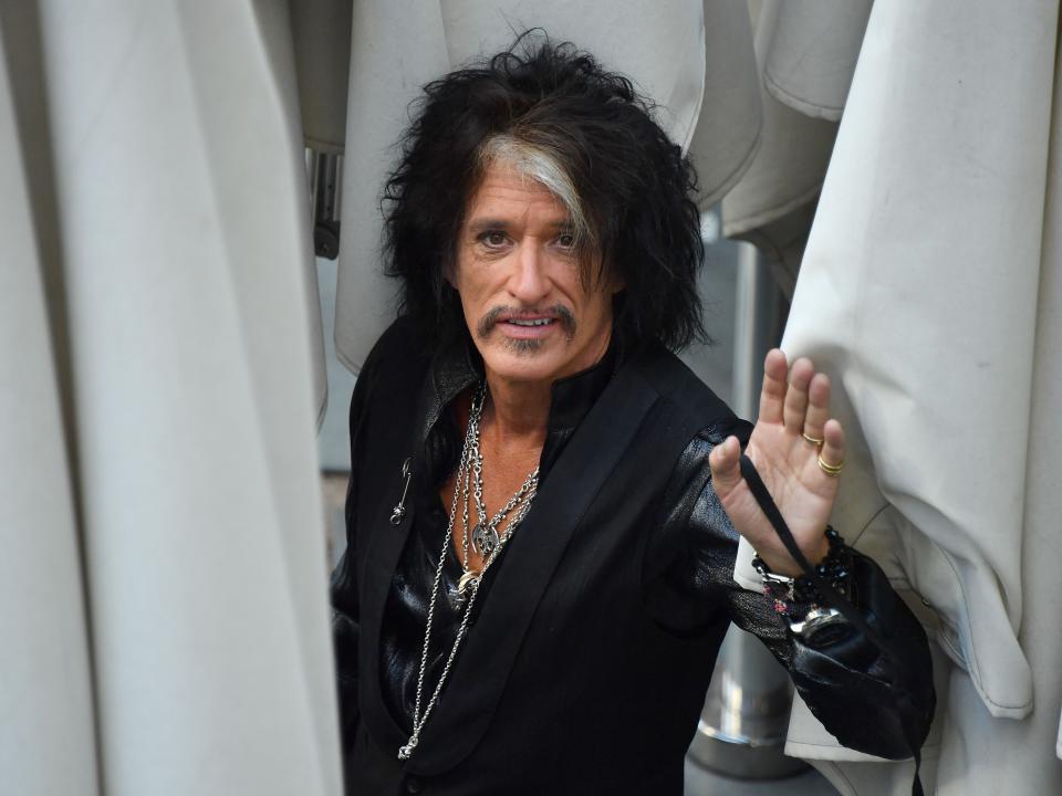 joe perry surrounded by white curtains, standing in the middle and waving and smiling towards the camera