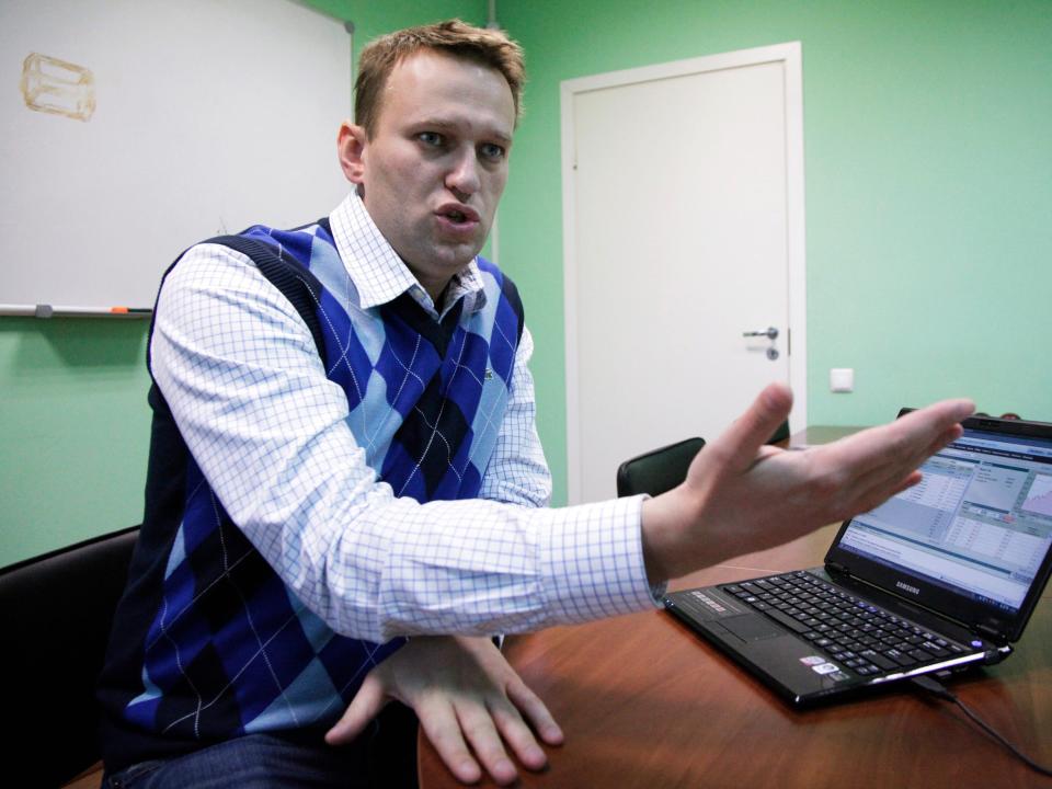 Alexei Navalny speaks and gestures with an outstretched arm while sitting at a desk in front of an open laptop.