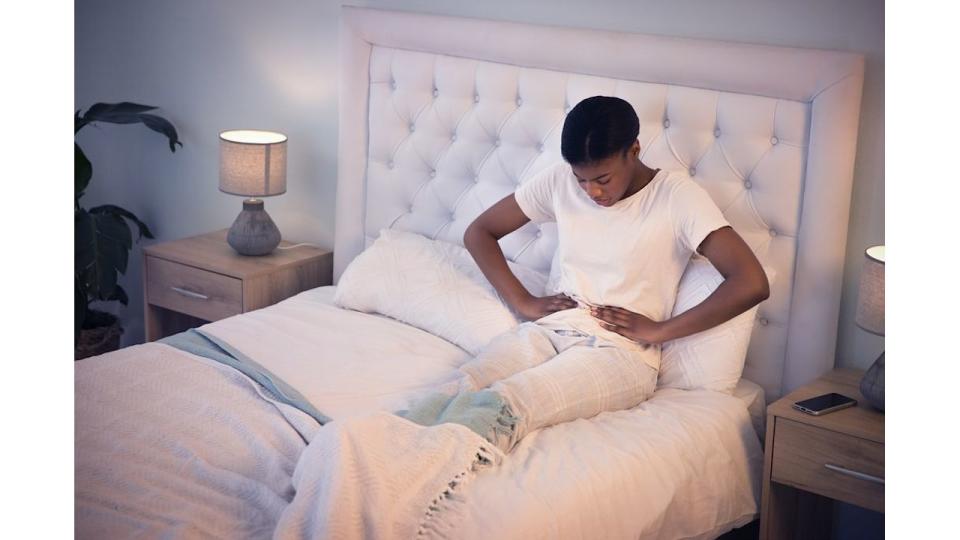 Pain, bed and woman with tummy cramps in her room holding her stomach for comfort. Tired, sick and African female person taking a nap with period ache for constipation or virus at her apartment.