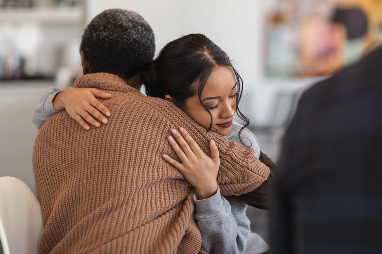 A young mixed-race woman hugs a mature adult black woman. They are sitting next to each other in a medical clinic. The two women are attending a group therapy session. They are showing support and kindness.