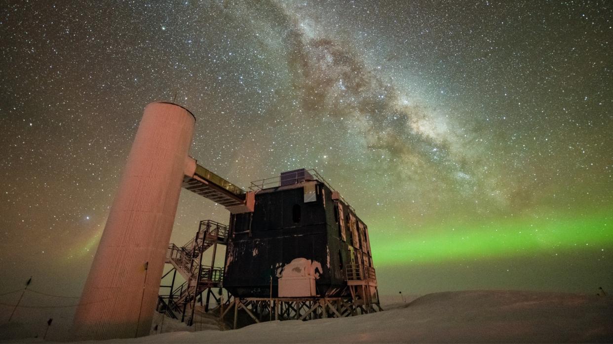  The IceCube Neutrino Observatory is seen under a starry night sky, with the Milky Way appearing over low auroras in the background. 