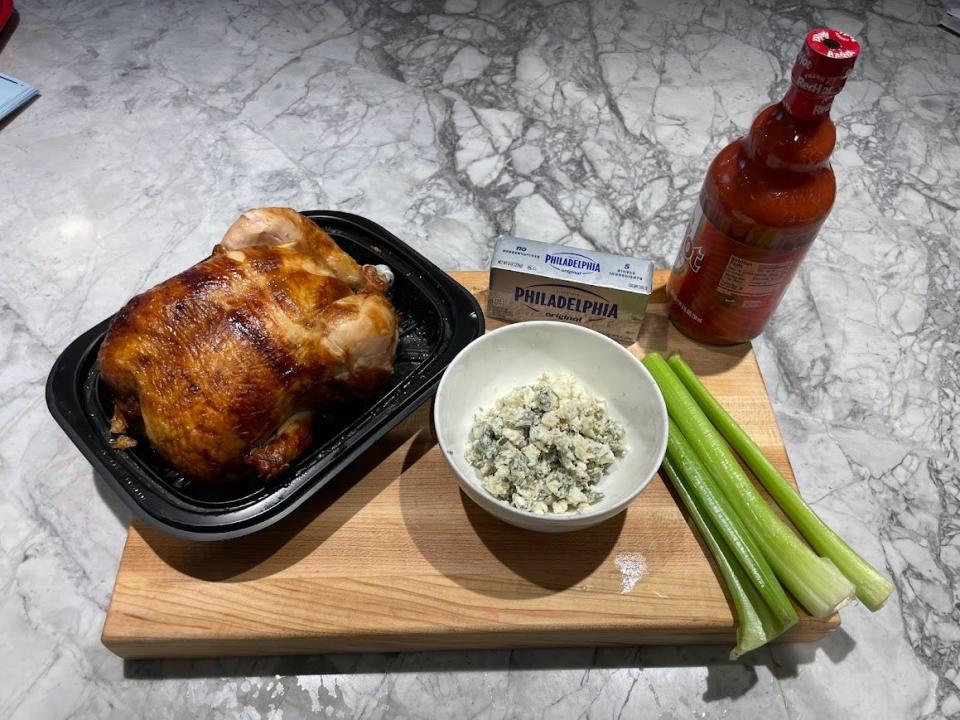 A cutting board with a rotisserie chicken, a bowl of cheese, a block of cream cheese, a bottle of hot sauce, and stalks of celery on it