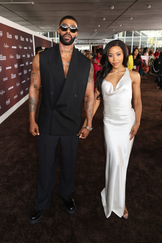Skyh Black, in a vest and pants, holds hands with KJ Smith, in a sleeveless gown