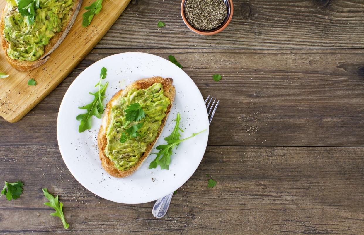 Avocado toast on a white plate on a rustic wooden table. The avocado is sprinkled with chopped parsley and black pepper, and served with fresh arugula. Space for copy on the right of the image. Stock photo.