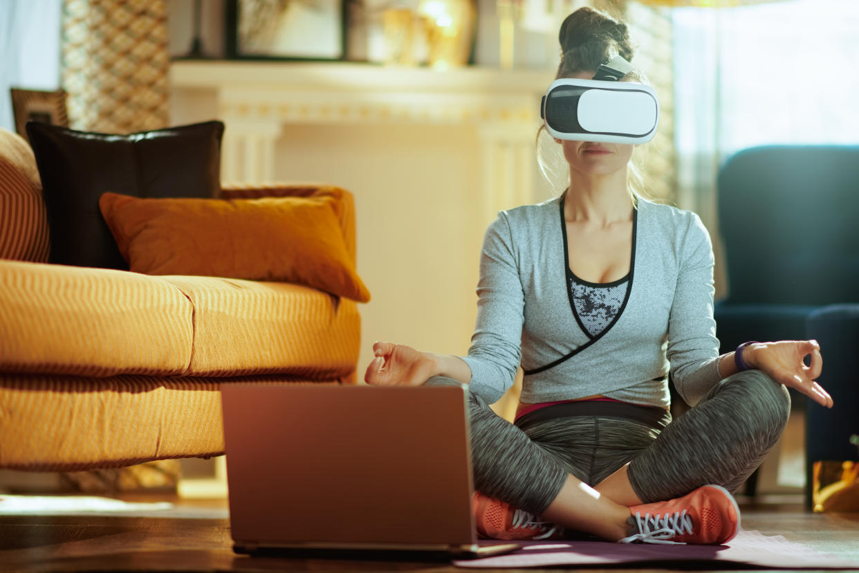 A relaxed healthy sports woman in sport clothes with laptop in VR gear meditating at modern home.