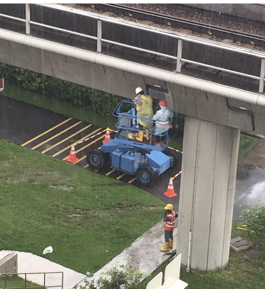 Two workers in safety gear on a blue lift platform under a raised railway track, with traffic cones nearby, performing maintenance or inspection work