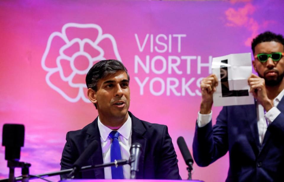 A man holds an L sign behind Rishi Sunak