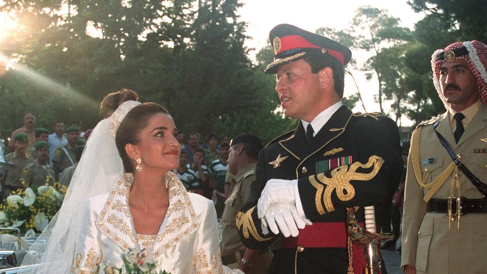 rania al yassin stands on a red carpet outside next to prince abdullah ii, she looks at him smiling and wearing an ornate ballgown style wedding dress with a long veil, she also holds a bouquet of greenery and white flowers, he wears a black military uniform with white gloves and a hat