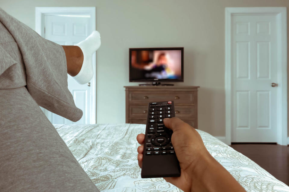 A woman's hand holding a TV remote, pointing it at the TV on the wall in the bedroom