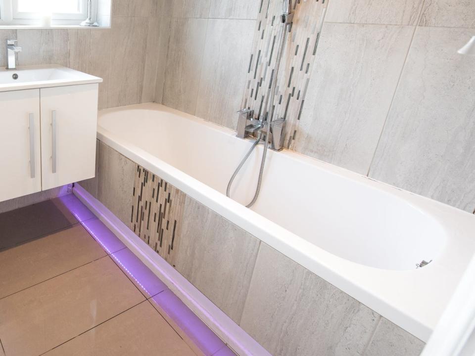 A bathroom with gray stone tile walls, a white toilet and floating sink countertop, and a tub with purple LED lights around the edge