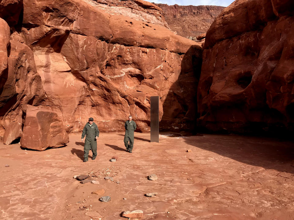 This Nov. 18, 2020 photo provided by the Utah Department of Public Safety shows Utah state workers walking near a metal monolith in the ground in a remote area of red rock in Utah. The smooth, tall structure was found during a helicopter survey of bighorn sheep in southeastern Utah, officials said Monday. State workers from the Utah Department of Public Safety and Division of Wildlife Resources spotted the gleaming object from the air and landed nearby to check it out. The exact location is so remote that officials are not revealing it publicly, worried that people might get lost or stranded trying to find it and need to be rescued. (Utah Department of Public Safety via AP)