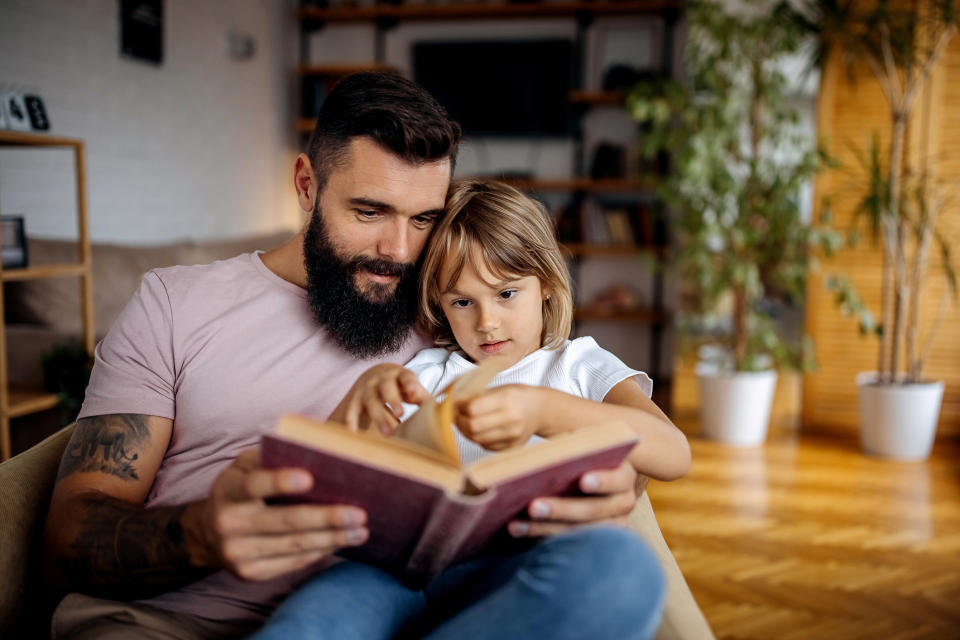 A bearded man sits with a young child on his lap, both are focused on reading a book together. The backdrop is a cozy living room with plants and bookshelves