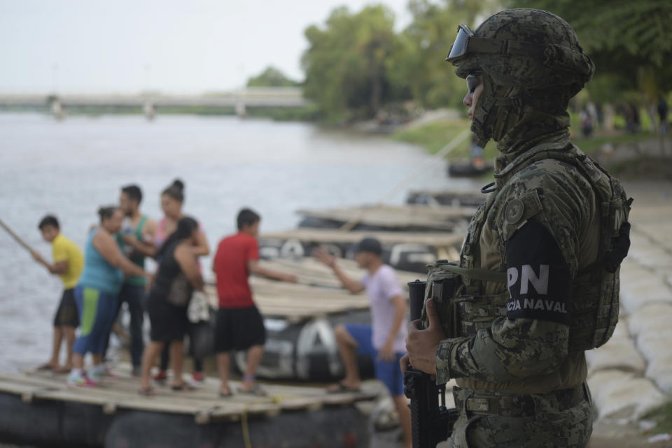 A Mexican marine stands guard on the Suchiate River, watching out for migrants crossing from Guatemala to Ciudad Hidalgo, as locals stand on a raft in Mexico, Sunday, June 16, 2019. Mexico faces heightened pressure from the U.S. to reduce the surge of mostly Central American migrants through its territory. (AP Photo/Idalia Rie)