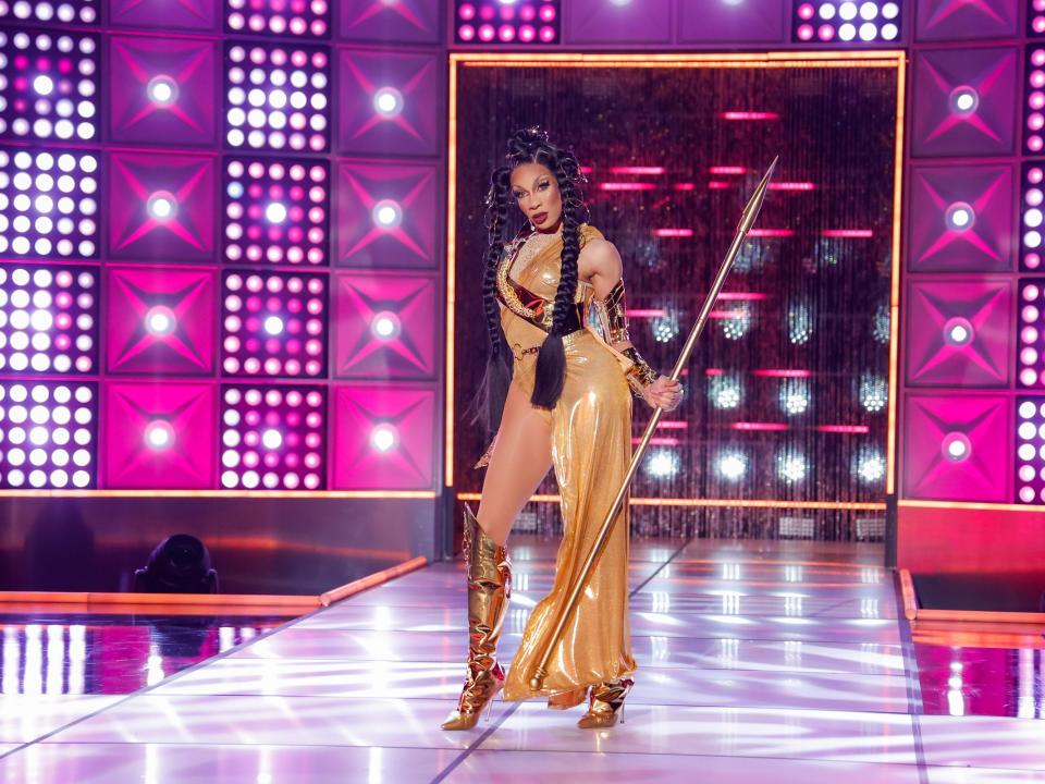 Robin Fierce poses on the runway in a gold gown with a gold spear and heeled boots in this still from episode 3 of "RuPaul's Drag Race" season 15.