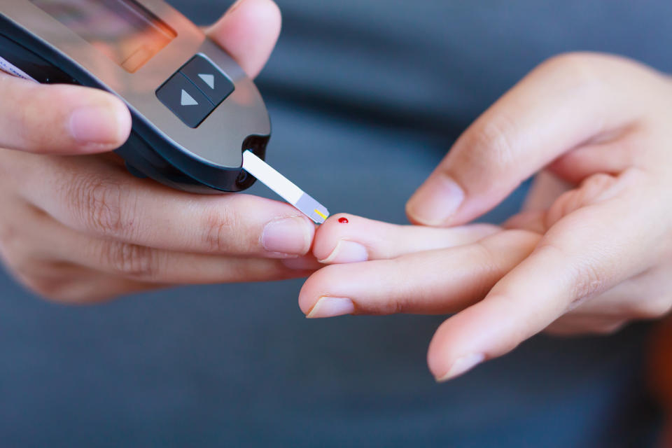 One hand, holding a glucose meter and strip, reaches to touch a drop of blood on the other.