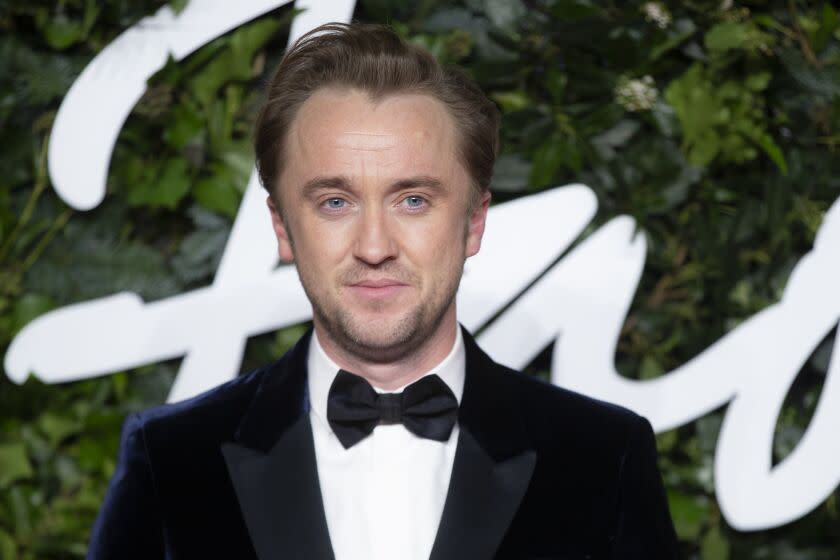 A man wearing a tuxedo stands in front of greenery