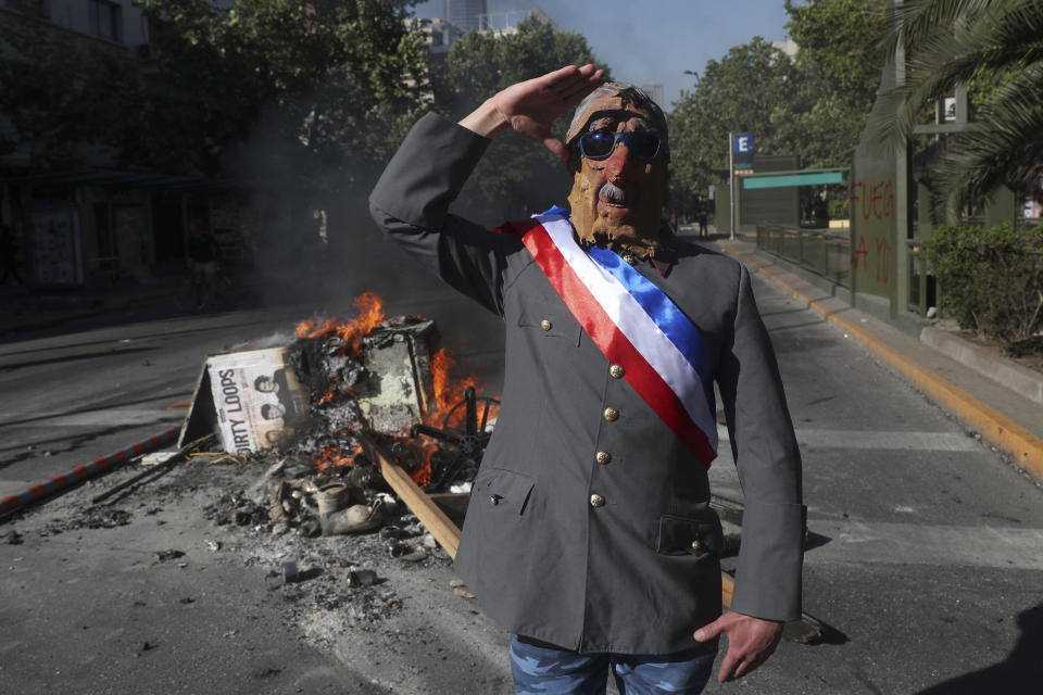 An anti-government demonstrator costumed as late military dictator Gen. Augusto Pinochet salutes in front of a burning barricade at a street in Santiago, Chile, Wednesday, Nov. 6, 2019. After nearly three weeks of anti-government protests many protesters are demanding a new constitution to replace the 1980 charter written under Gen. Augusto Pinochet's 1973-1990 military dictatorship. (AP Photo/Esteban Felix)
