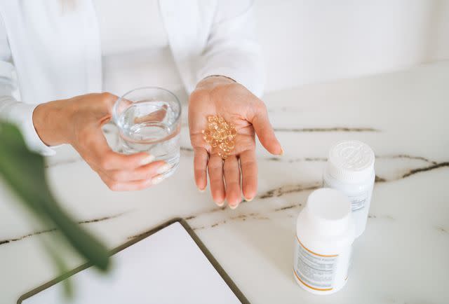 <p>Galina Zhigalova / Getty Images</p> Female healthcare provider in white sweater with omega 3, vitamin D capsules in one hand and a glass of water in the other.