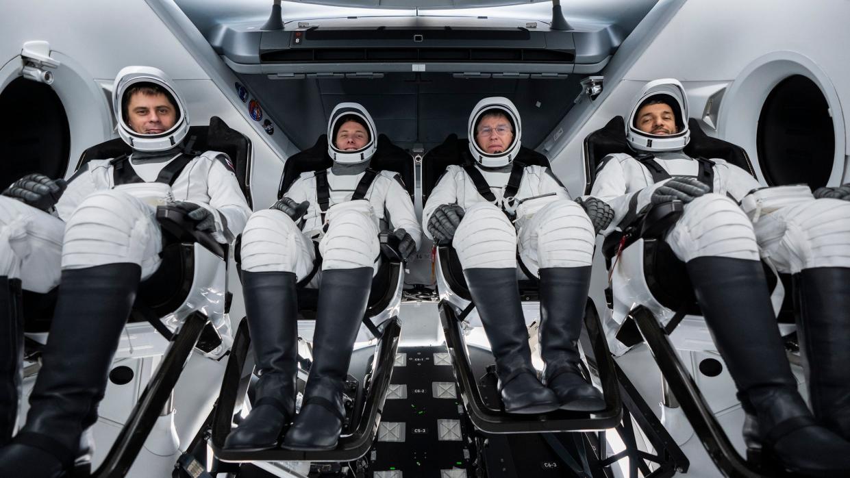  four spacesuited astronauts sitting side by side in a spacecraft with visors up and seatbelts on 