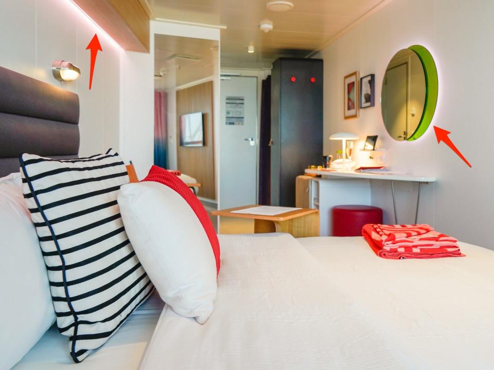 A bed with white sheets and a red banket and throw pilloe with a desk and a mirror behind it. Two red arrows point to mood lighting behind a mirror and above the bed.