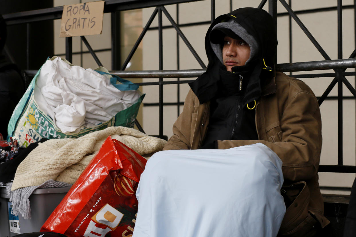 A young man in a hooded jacket with a blue sheet over his legs sits on scaffolding beside half a dozen bags and plastic buckets packed with belongings.