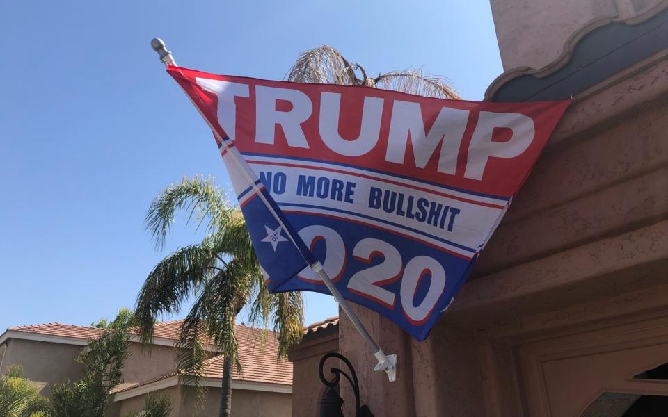 A red and white flag reading "TRUMP 2020: No More Bullshit" flutters in the breeze on a pole in front of a tan-coloured garage, with palm trees in the background - Laurence Dodds/Telegraph