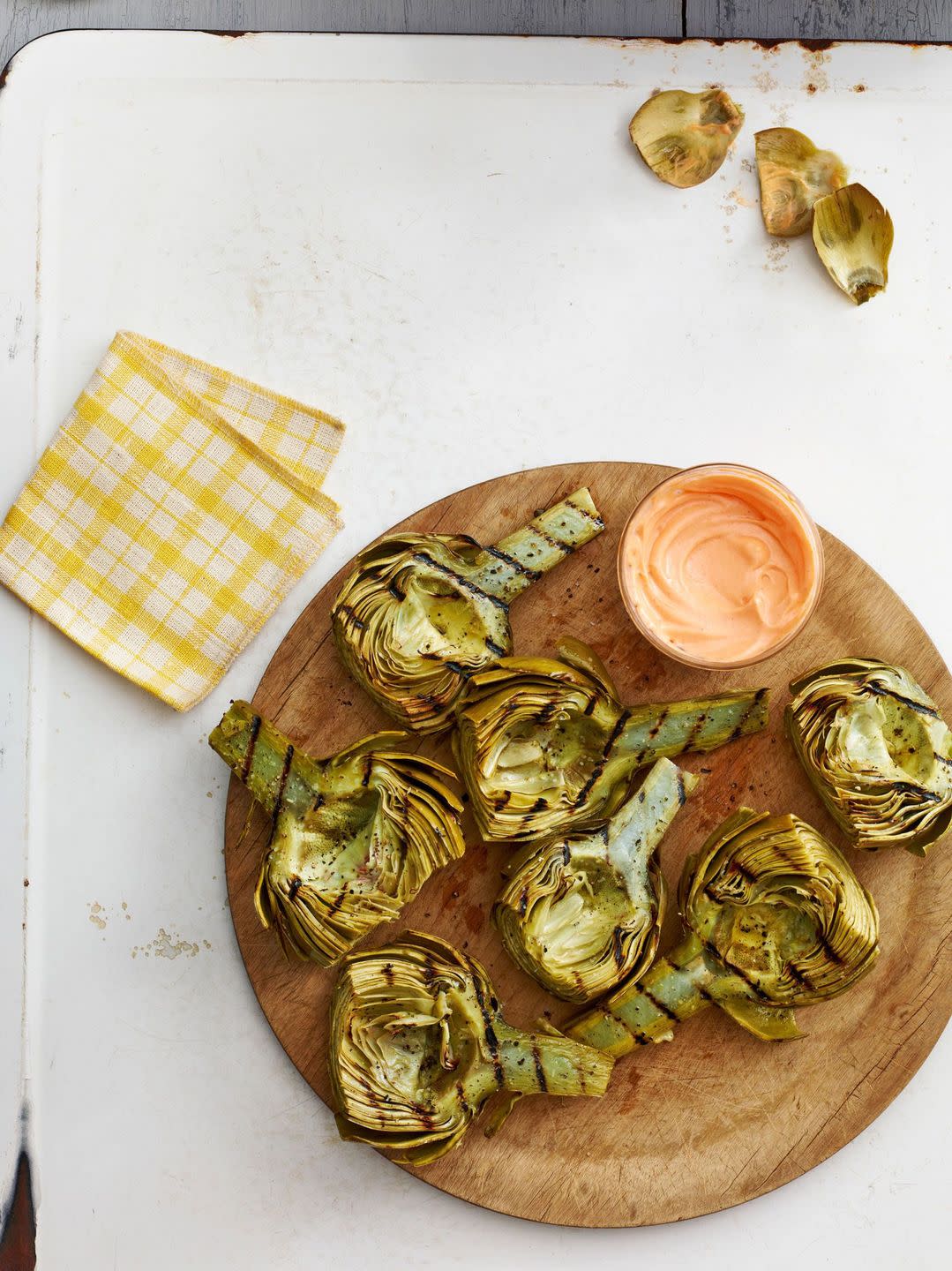 grilled artichokes on a wooden plate with a small bowl of harissa honey dip