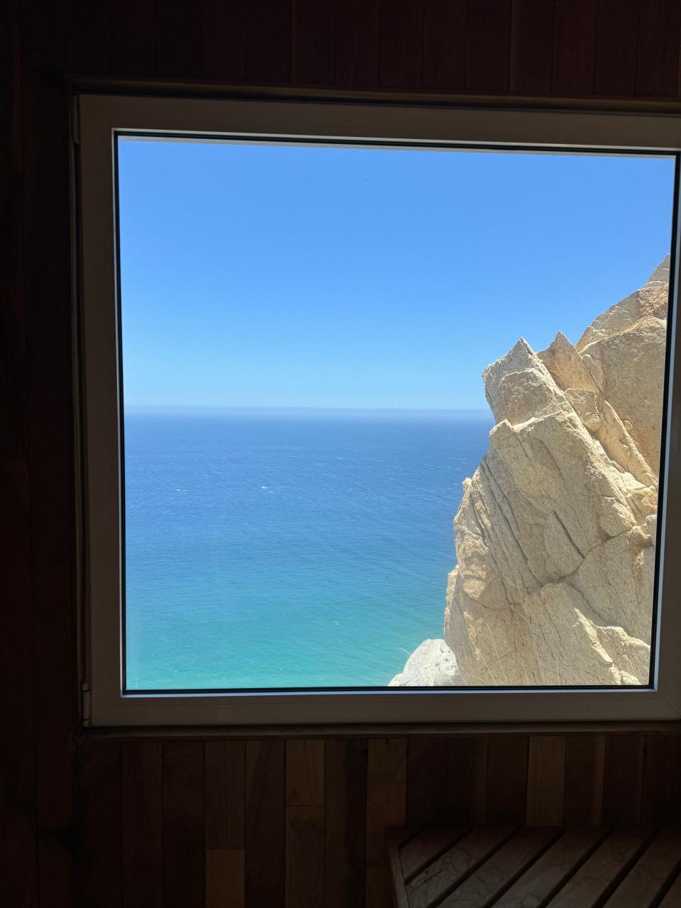 A window view shows a close-up of rocky cliffs above a clear, blue ocean under a bright sky. Wood paneling is partially visible around the window frame