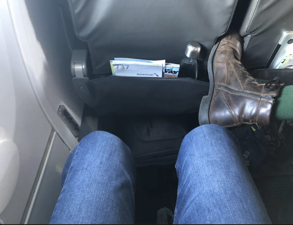 Person's legs with jeans in an airplane seat, safety card in seatback pocket, and a brown boot on the right