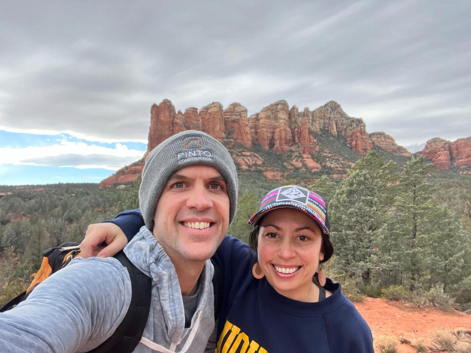 A man and a woman take a selfie while hiking in front of red rock formations.